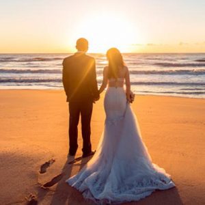 Newlywed husband and wife holding hands on a beach as the sun sets.