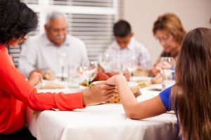 Hispanic family sitting at the dinner table, holding hands in prayer.