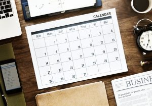 calendar in middle of clock, phone, newspaper and planner on a wooden table