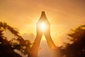 Person's prayer hands with image of sunlight and clouds overlayed.